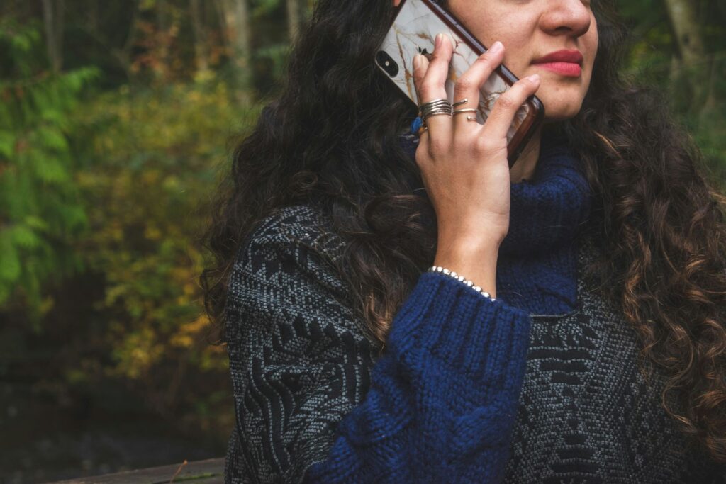 Close-up of a woman holding a phone to her ear, listening intently while standing outdoors in a wooded area, wearing a cozy blue sweater and layered rings.