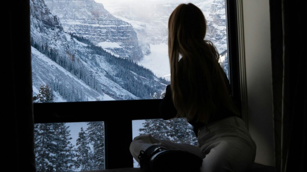 A woman sits quietly by a large window overlooking a snowy mountain valley, reflecting on peace, possibility, and the meaning of the holiday season.