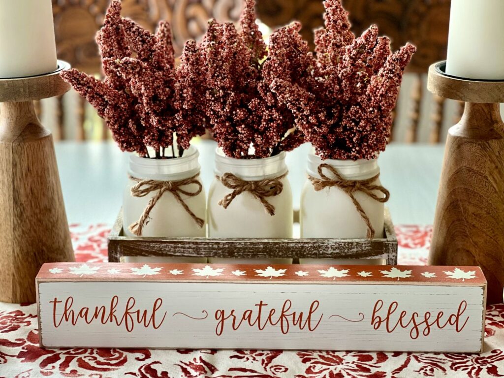 A cozy Thanksgiving table display with mason jars filled with rustic flowers and a wooden sign reading “thankful • grateful • blessed,” symbolizing quiet gratitude and simple holiday reflection.